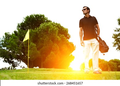 Full length portrait of stylish golfer in glasses standing on golf course with amazing flare sunset light on background, handsome man with golf bag on shoulder standing on golf course, filtered image