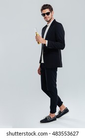 Full Length Portrait Of A Stylish Young Man In Black Suit And Sunglasses Holding Beer Bottle Over Grey Background