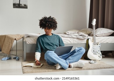Full length portrait of teenage African-American student sitting on floor at home or in college dorm and studying , copy space