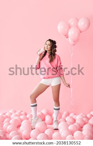 Full length portrait of trendy young woman drinking milk shake over pink balloons background. Creative, celebration, pop art concept