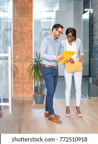 Full Length Portrait Of Two Business People With Yellow Folders Talking In Office Hall.