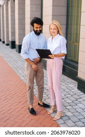 Full Length Portrait Vertical Portrait Of Attractive Young Blonde Business Woman Explaining Something On Clipboard To Male Business Partner, Standing In City Street.