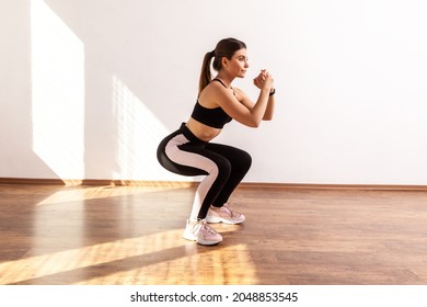 Full Length Portrait Of Woman Doing Split Or Lunge Squat Exercise At Home Or Fitness Gym, Wearing Black Sports Top And Tights. Indoor Studio Shot Illuminated By Sunlight From Window.