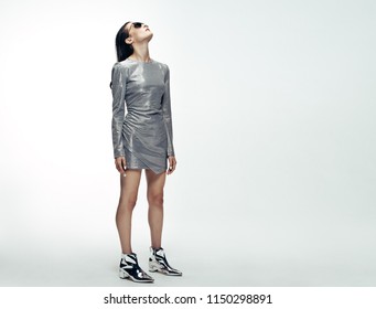 Full length portrait of woman in silver dress looking upwards while standing over grey background. Female model in futuristic style. - Shutterstock ID 1150298891