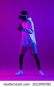 Full Length Portrait Of Young African Man Wearing VR Glass Headset Standing And Punching In Futuristic Dark Purple Cyberpunk Neon Light Background