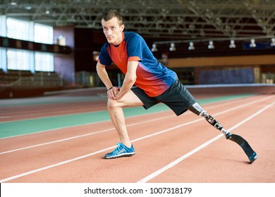 Full length portrait of young amputee sportsman  warming up before running practice in modern gym stretching legs on track, copy space
