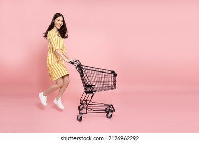 Full length portrait of young Asian woman jumping and pushing an empty shopping cart or shopping trolley isolated on pink background