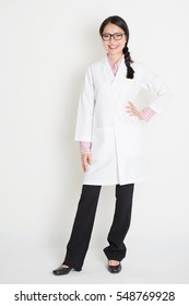 Full Length Portrait Of Young Asian Female Scientist With Lab Coat Standing On Plain Background.