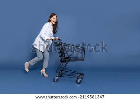 Full length portrait of young Asian woman pushing an empty shopping cart or shopping trolley isolated on deep blue background