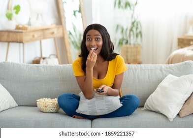 Full length portrait of young black woman with remote control watching TV and eating popcorn on sofa at home. Positive African American lady enjoying good movie or program on weekend