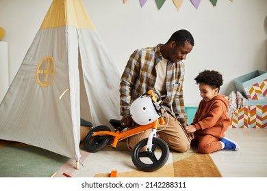 Full Length Portrait Of Young Black Father Playing With Toddler Son Indoors And Teaching Him To Ride First Balance Bike