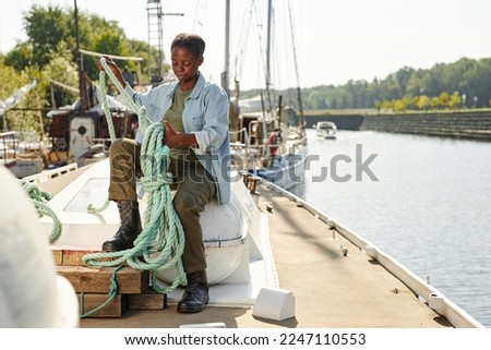 Full length portrait of young black woman tying rope on boat in docks lit by sunlight, copy space