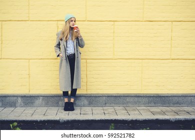 Full length portrait of young blonde female in trendy casual outfit drinking coffee outdoors standing on urban setting background copy space area for advertising while waiting for best friends meeting