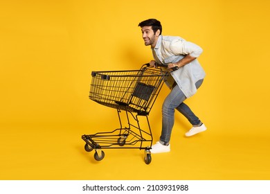Full length portrait of young Caucasian man pushing an empty shopping cart or shopping trolley isolated on yellow background
