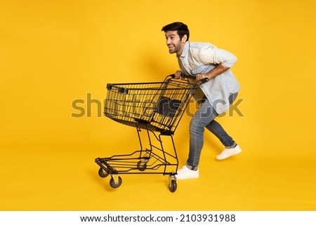 Full length portrait of young Caucasian man pushing an empty shopping cart or shopping trolley isolated on yellow background