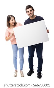 Full Length Portrait Of A Young Couple Holding A White Sign While One Of Them Looks At It In A Studio