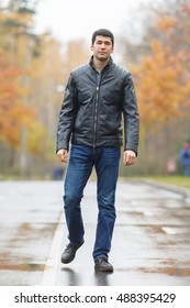 Full Length Portrait Of Young Dark-haired Man In Black Jacket On Avenue In Park, Walking