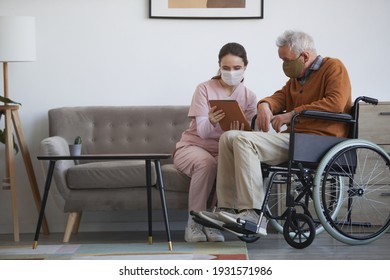 Full length portrait of young female nurse assisting senior man in wheelchair using digital tablet at retirement home, both wearing masks, copy space