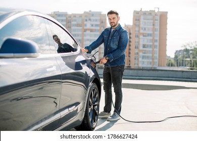 Full length portrait of young handsome bearded man in casual wear, standing at the charging station and holding a plug of the charger for an electric car. Eco electric car concept