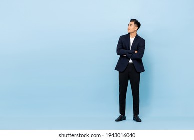 Full length portrait of young handsome southeast Asian businessman with arms crossed looking up sideway to copy space on light blue studio background