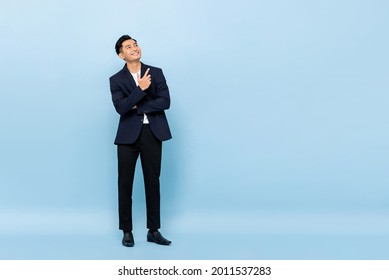 Full length portrait of young handsome southeast Asian businessman looking up and pointing to copy space on light blue studio background - Shutterstock ID 2011537283