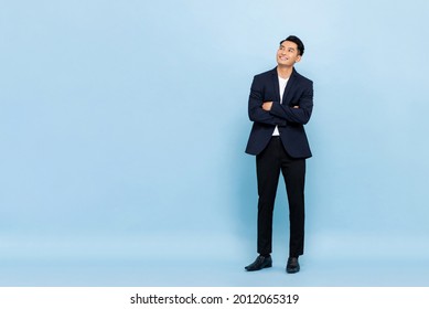 Full length portrait of young handsome southeast Asian businessman with arms crossed looking up to copy space on light blue studio background