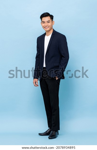 Full length portrait of young handsome\
southeast Asian millenial businessman looking at camera on light\
blue studio background