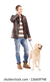 Full Length Portrait Of A Young Man Walking A Dog And Talking On A Mobile Phone, Isolated On White Background