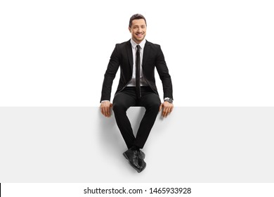 Full Length Portrait Of A Young Man In A Suit Sitting On A Panel And Smiling Isolated On White Background
