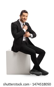 Full Length Portrait Of A Young Man In A Suit Sitting And Fixing His Tie Isolated On White Background