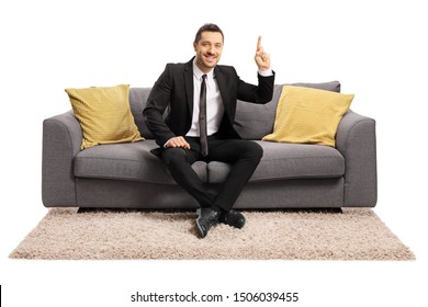 Full length portrait of a young man in a black suit sitting on sofa and pointing up isolated on white background
