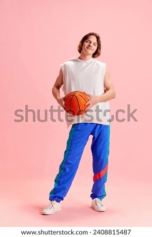 Full length portrait of young man dressed in retro sport uniform and holding basketball ball against pastel pink background. Concept of active lifestyle, sport and recreation, hobby, fashion.