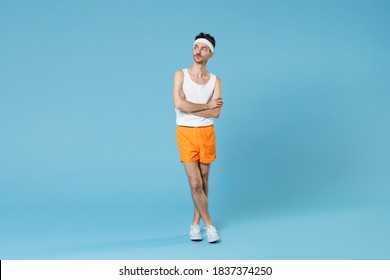 Full Length Portrait Of Young Sporty Fitness Man With Skinny Body Sportsman In White Headband Shirt Shorts Holding Hands Crossed Isolated On Blue Background. Workout Gym Sport Motivation Concept