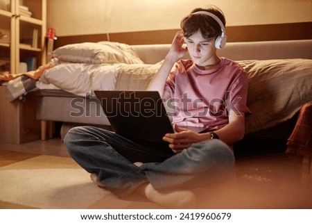Full length portrait of young teenage boy using laptop while sitting on floor at night with legs crossed and wearing wireless headphones