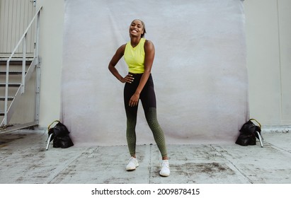 Full Length Portrait Of Young Woman In Exercise Outfit Smiling While Relaxing On Rooftop. African Athlete Woman With Fit Body Taking Break Post Workout On Building Terrace.