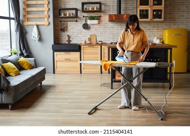 Full length portrait of young woman ironing clothes at home and doing household chores on weekend, copy space - Powered by Shutterstock