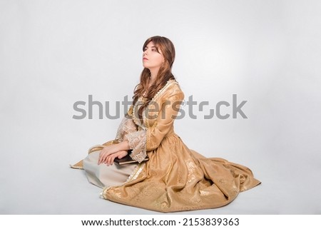 Full length portrait of a young woman in a puffy gold dress in the rococo era, posing while sitting with a book in her hands, isolated on a white background. The girl is reading a book.
