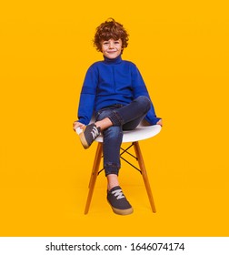 Full length positive child model in stylish outfit smiling for camera and sitting on chair against yellow background