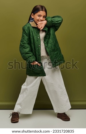 Full length of positive preadolescent girl with dyed hair looking at camera while posing in autumn outfit and knitted sweater on green background, modern fall fashion for preteens concept