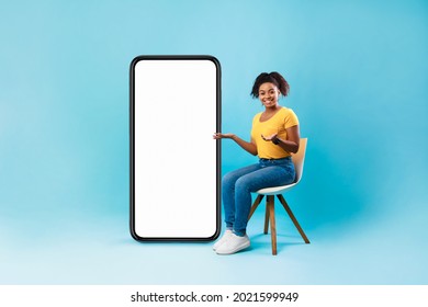 Full length of pretty Afro lady sitting on chair next to big blank smartphone with mockup for mobile app or website design, blue studio background. Space for your online advertisement