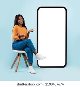 Full length of pretty black lady sitting on chair next to big blank smartphone with mockup for mobile app or website design, blue studio background, showing copy space for your online advertisement