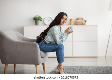 Full length of pretty Indian lady sitting in armchair with cup of hot coffee at home, copy space. Millennial woman enjoying aromatic beverage on cool day, relaxing with her favorite warm drink - Powered by Shutterstock