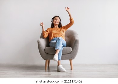 Full length of pretty young Asian woman listening to music in headphones, dancing while sitting in armchair against white studio wall. Lovely millennial female enjoying her favorite song - Shutterstock ID 2111171462