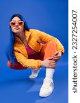 full length of pretty young female model with blue hair and trendy sunglasses sitting on haunches on blue background, generation z, rebel style, individualism, modern fashion, trendy accessory