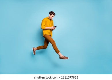 Full Length Profile Photo Of Funny Guy Jumping High Holding Telephone Millennial Writing Email Walking Down Street Wear Yellow Shirt Pants Isolated Blue Color Background