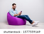 Full length profile photo of positive guy sitting comfy violet armchair holding telephone chatting friends wear specs casual denim outfit isolated grey color background