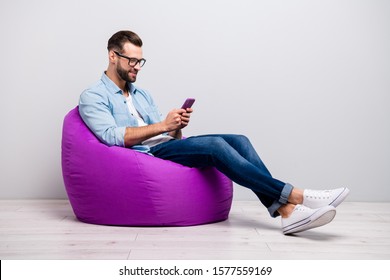 Full length profile photo of positive guy sitting comfy violet armchair holding telephone chatting friends wear specs casual denim outfit isolated grey color background
