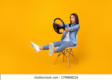 Full length profile photo of pretty funny lady good mood sit chair spread legs hold steering wheel riding imagine car rushing wear casual denim shirt shoes isolated yellow color background