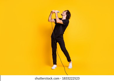 Full Length Profile Photo Of Young Man Hold Mic Sing Open Mouth Wear Black T-shirt Pants White Sneakers Glasses Isolated Yellow Color Background