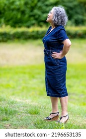 Full Length Profile Portrait Of Smiling Mature Woman Standing Against Blurred Green Background, Looking Up, Wearing Denim Dress, Platform Sandals, Short Flowing Gray Black Hair, Enjoying Sunny Day